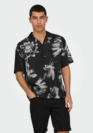 Camisa negra de manga corta con estampado floral que presenta contornos blancos de grandes flores y hojas, con botones en la parte delantera y un corte relajado. Combinada con pantalones cortos negros.