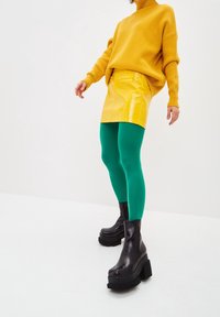 Maglione giallo a coste, mini gonna gialla lucida, leggings verdi e stivaletti neri chunky alla caviglia. Focalizzarsi su colori vivaci e contrasti di materiali.