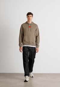 Felpa khaki con tasca frontale e etichetta rossa, abbinata a jeans in denim scuro. Maglietta bianca visibile all'orlo, indossata con scarpe da ginnastica bianche.