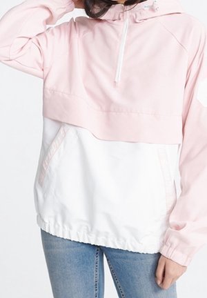 Femme portant une veste coupe-vent bicolore rose et blanche avec une fermeture éclair à mi-chemin, des poches avant et un jean bleu.