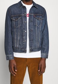 Chaqueta de mezclilla azul oscuro con dos bolsillos en el pecho, botones plateados y detalles de costura sutiles. Llevada sobre una camiseta blanca y pantalones de pana marrón.