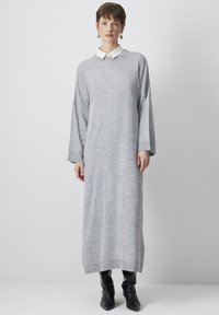 Rochie gri, cu mânecă lungă, confecționată dintr-un material tricotat moale, având un decolteu rotund și o siluetă dreaptă, purtată peste cizme negre până la genunchi.