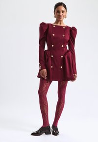 Burgundské šaty s nafouknutými rukávy a zlatými knoflíky, strukturovaná silueta, detail skladaného pasu, kombinované s burgundskými krajkovými punčochami a černými botami.