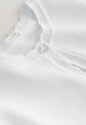 Haut en coton blanc avec un col à volants, comportant un bouton décoratif et une étiquette indiquant "NEXT, FABRIQUÉ AU BANGLADESH."