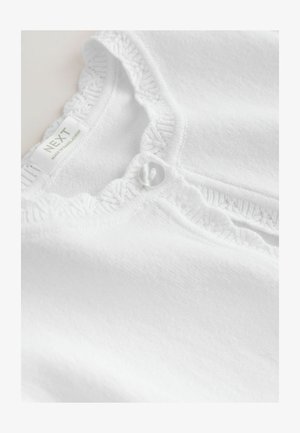 Top din bumbac alb cu guler volan, având un singur nasture decorativ și un etichetă care spune "NEXT, MADE IN BANGLADESH."