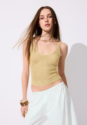 Ung kvinde med langt brunt hår iført en beige strikket ærmeløs top, hvide bukser og chunky armbånd, poserer mod en ensfarvet baggrund.