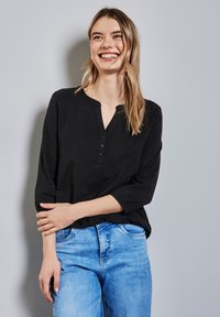 Schwarzes Langarm-Henley-Oberteil mit drei Knöpfen am Ausschnitt, kombiniert mit hellblauen Denim-Jeans. Glatte Textur und lockere Passform.