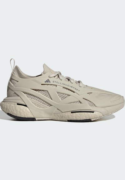 Zapatilla atlética beige con una parte superior de malla flexible, diseño en capas distintivo, suela texturizada y acentos mínimos. Presenta un robusto tirador en el talón.