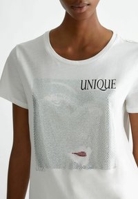 T-shirt bianca a maniche corte con una grafica decorata con strass, la parola "UNIQUE" in nero e un accento rosso nel design.