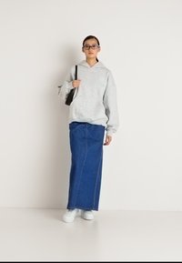 Mulher jovem a usar óculos, hoodie cinza, saia de ganga azul longa, ténis brancos e a carregar uma mala preta de ombro, em pé contra um fundo branco.