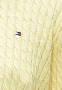 Pull en maille torsadée jaune clair avec un motif texturé et un petit écusson logo présentant des accents rouges, blancs et bleus.
