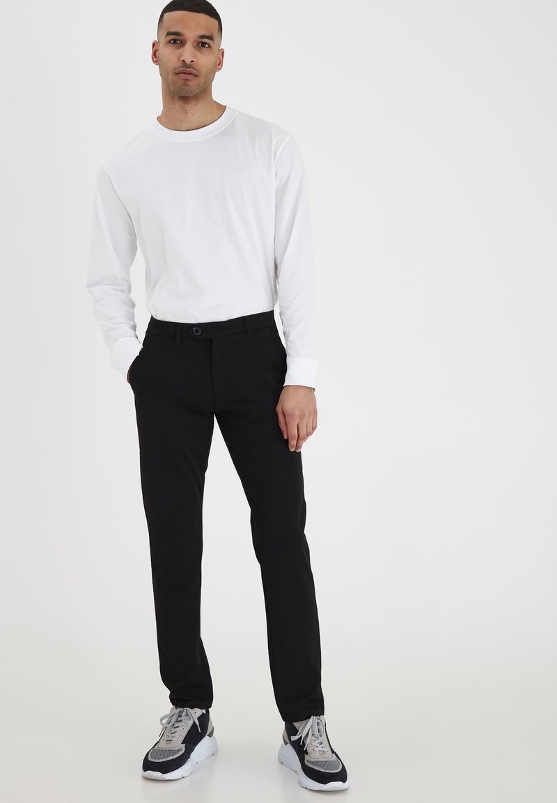 Črne krojene hlače, slim fit z gladko teksturo, v kombinaciji z belim dolgimi rokavi. Teniske imajo sivo in mornarsko modre detajle.