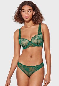 Sujetador verde de encaje y tanga a juego con patrones florales, bordes con forma de concha y detalles de malla transparente. Tirantes ajustables en el sujetador.
