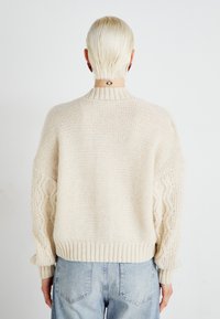 Kremet chunky strikkegenser med ribbestrikket krage og mansjetter, samt teksturerte kabelstrikkede detaljer på ermerne. Ensfarget bak og løs passform.