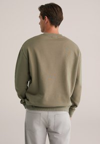 Olivno zelena majica s okruglim izrezom i spuštenim ramenima, koja ima suptilan teksturiran uzorak na leđima, u kombinaciji s svijetlosivim hlačama.