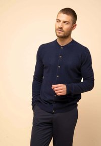 Cardigan azul marino en suave tejido de punto, con cuello redondo, cierre de botones en la parte delantera y puños acanalados, combinado con pantalones oscuros.