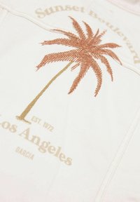 Bílá látka s designem palmového stromu v bronzové výšivce. Text uvádí "Sunset Boulevard Los Angeles EST. 1972" ve zlaté barvě.
