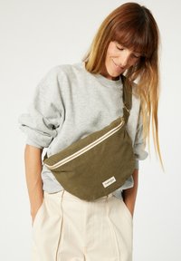 Sac bandoulière en toile vert olive avec une fermeture éclair blanche, texture lisse et une petite étiquette de logo. Forme incurvée et bandoulière réglable.
