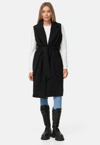Schwarzer ärmelloser Mantel mit einem Gürtel in der Taille, breiten Revers und strukturiertem Finish, kombiniert mit einem weißen Pullover und blauen Jeans.