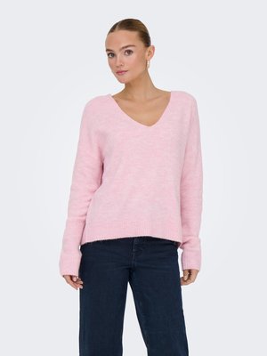 Femme portant un pull rose clair à col en V avec des manches longues et un jean bleu foncé, debout devant un fond clair uni.