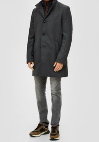 Cappotto grigio in misto lana con colletto a scollo, due bottoni, tasche laterali, indossato sopra una giacca imbottita nera, abbinato a jeans grigi e sneakers.