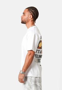 Hombre con cabello trenzado, vestido con una camiseta blanca con gráfico, pantalones claros con patrón, un reloj y un collar de cuentas, de pie de perfil contra un fondo blanco.