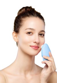 Dispositif de nettoyage facial en silicone bleu tenu contre la joue d'une personne. Surface lisse avec des nervures texturées, conçu pour les soins de la peau.