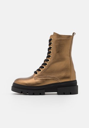 Botte de combat en cuir bronze métallisé avec semelle épaisse en caoutchouc noir et col rembourré à la cheville sur fond blanc.