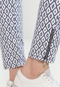 Pantalones blancos con un patrón de diamantes azul marino, con un detalle de cremallera lateral en el tobillo, combinados con zapatillas deportivas blancas.