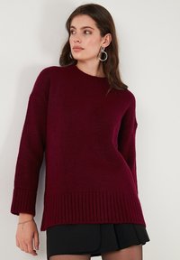 Burgundy színű pulóver bordázott textúrával, kerek nyakkivágással és esésvállú dizájnnal. Hosszú ujjak jellemzik, bordázott aljjal a részletek fokozása érdekében.