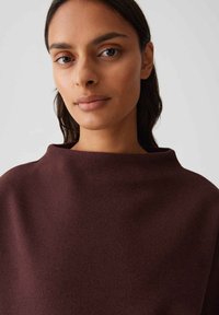 Vrouw met donker haar, gekleed in een hoge hals, lange mouwen, donkerbruine top tegen een effen lichte achtergrond.