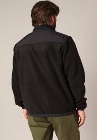 Zwarte fleecjas met een hoge kraag en een contrasterend stoffen paneel op de schouders. Geribde manchetten en onderrand. Lichtgewicht textuur.