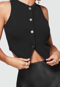 Schwarzes, geripptes, ärmelloses Cropped-Top mit hohem Kragen, vorderer Knopfverschluss und schrägem Saum. Auffällige Textur und glatte Knopf-Details.