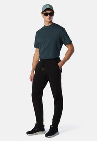 Uomo con una camicia a maniche corte di colore verde scuro, pantaloni jogger neri, sneakers nere, berretto verde e occhiali da sole, in piedi con una mano in tasca.