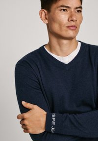 Námořnická modrá svetr s výstřihem do V z měkké tkaniny, opatřený pružnými manžetami s textem "Pepe Jeans". Design je jednoduchý s přiléhavým střihem.