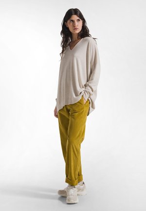 Femme debout portant un pull beige ample, un pantalon en velours côtelé jaune moutarde et des baskets claires sur un fond blanc uni.