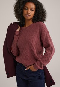 Pull en mailles texturé bordeaux avec un motif géométrique, col rond et manches longues, associé à un manteau bordeaux foncé. Cheveux bouclés visibles.