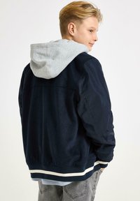 Marineblaue Bomberjacke mit grauem Kapuzen. Verfügt über einen gerippten, weiß gestreiften Saum und ein strukturiertes Material am Körper und an den Ärmeln. Lässiger Schnitt.