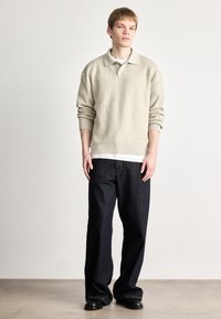 Pulover bej cu dungi, cu guler polo, purtat peste o cămașă albă; asociat cu blugi largi de culoare albastru închis și pantofi negri.