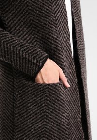 Dlouhý, tmavě hnědý kardigan s vzorem rybí kosti, texturovaný vlněný materiál, otevřený přední díl a boční kapsy.