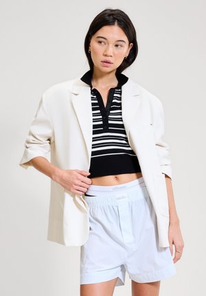 Kobieta ubrana w biały oversize'owy blezer, czarno-białą paskowaną krótką koszulę i jasnoniebieskie szorty, pozująca na jednolitym tle.