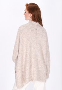 Beige, oversize kötött pulóver magas gallérral és texturált felülettel. Hátul varrás és diszkrét logó részlet található a gallérnál.