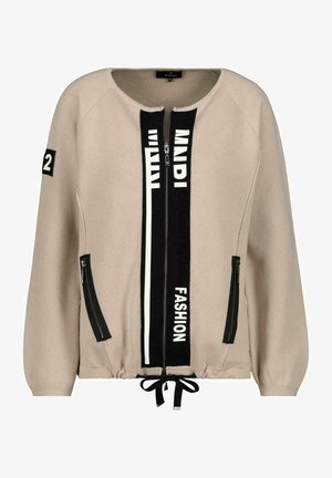 Beige ritsjack gemaakt van gestructureerde stof, met zwarte accenten en witte tekst, een elastische tailleband en twee zijzakken met rits.