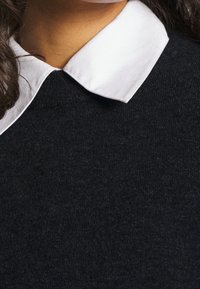 Camicia bianca con colletto sotto un maglione nero strutturato, con capelli scuri e mossi parzialmente visibili sul lato sinistro.