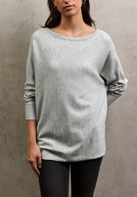 Světle šedý oversized svetr s širokým výstřihem a dlouhými rukávy, vyrobený z měkké látky. V kombinaci s tmavými džíny na neutrálním pozadí.