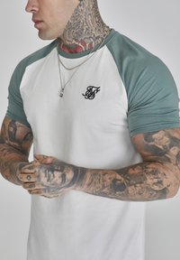 Tricou scurt cu mânecă albă și mâneci raglan de culoare verde deschis, având un logo brodat negru și tatuaje texturate pe antebrațe.