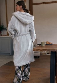 Lichtgrijze badjas met capuchon van gestructureerde stof, tailleband en lange mouwen, gecombineerd met geruite broek. Houten tafel met een mok en croissants.