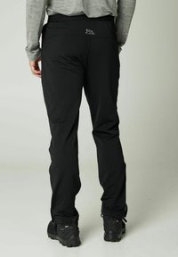 Schwarze Outdoor-Hose mit glatter Textur und elastischem Bund. Mit einem Berglogo auf der Rückseite versehen, kombiniert mit robustem Schuhwerk.