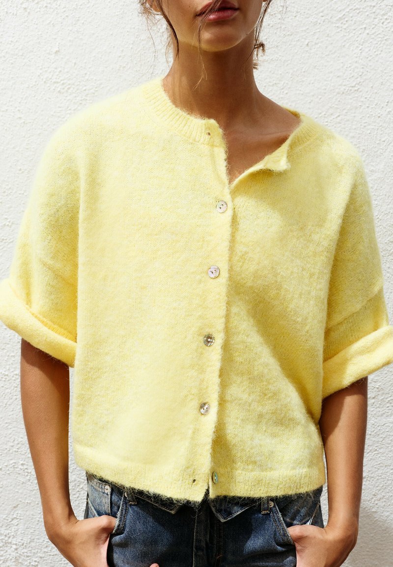 Femme portant un pull jaune ample et duveteux boutonné avec des manches courtes retroussées et un jean bleu, debout devant un mur blanc texturé.