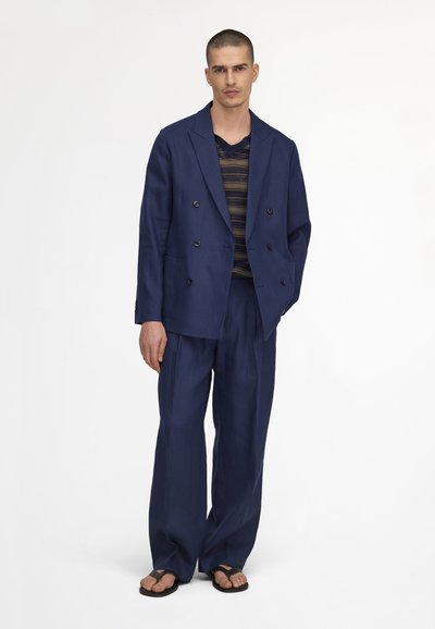 Homme debout portant un costume croisé bleu marine ample, un haut en tricot rayé et des tongs noires sur fond blanc.
